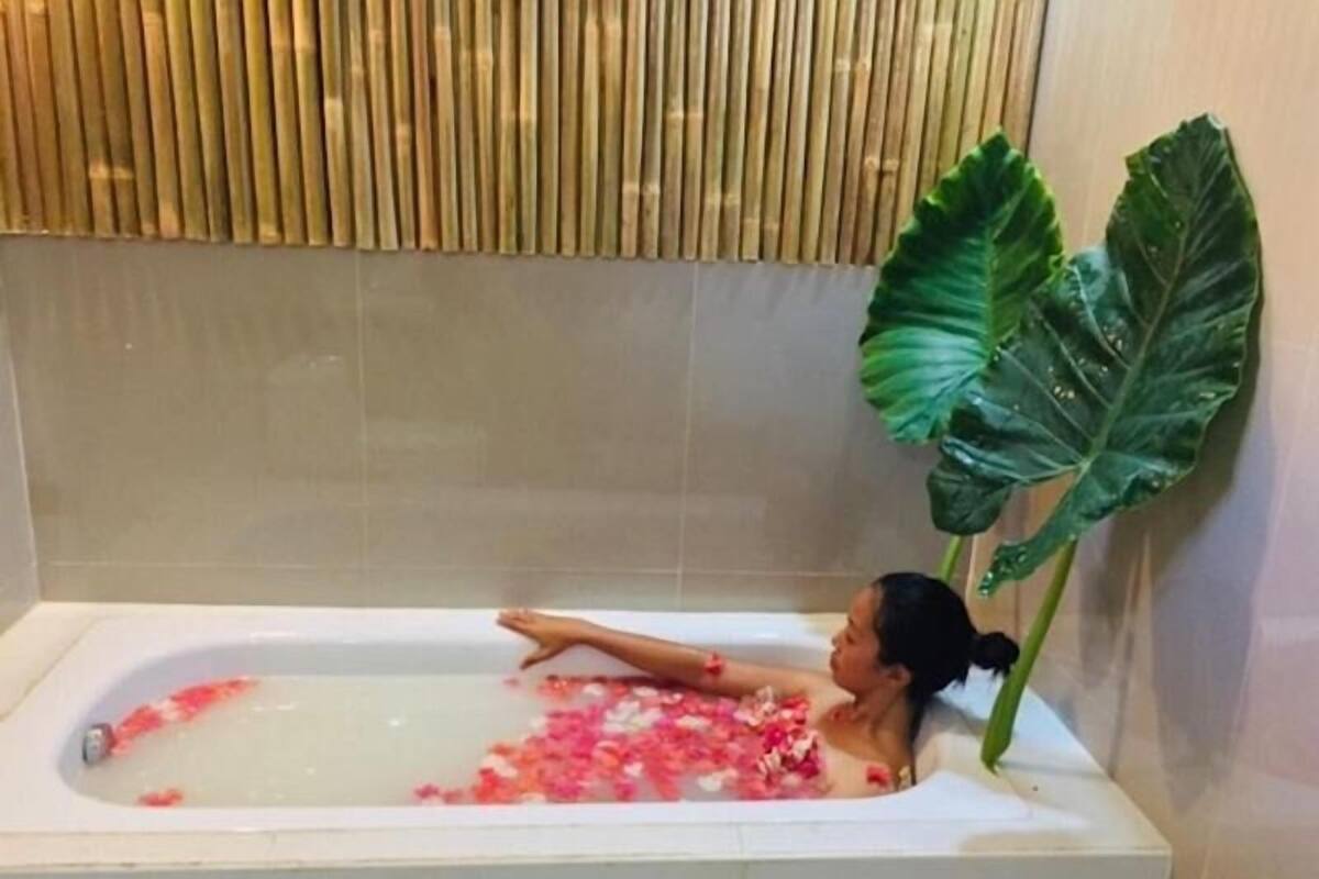 A bathtub with water and flower petals, a bamboo wall, and a large green plant.