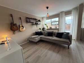 Ein Wohnzimmer mit einer Eckcouch, zwei Gitarren an der Wand, einem Bücherregal, einer Topfpflanze und einem großen Fenster mit Vorhängen.