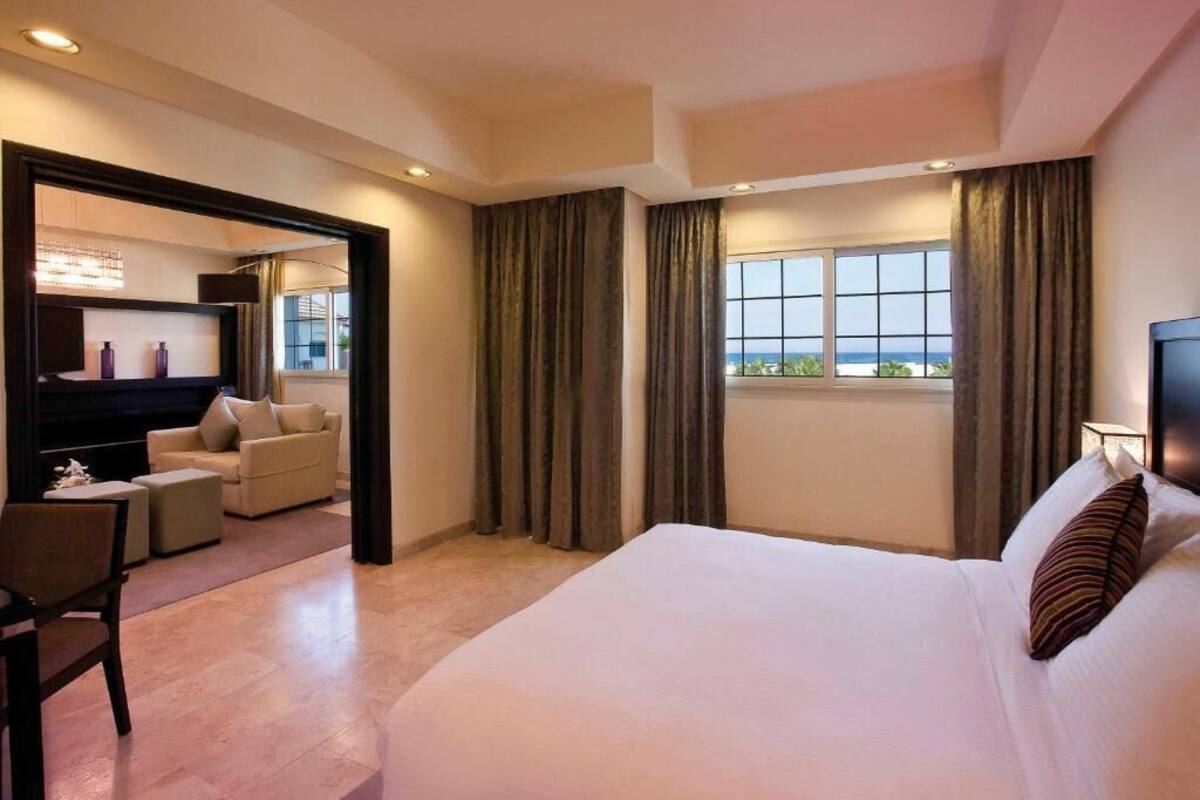 Ein Hotelzimmer mit einem großen Bett, einer Sitzecke mit einer Couch und Sesseln, einem Fernseher und einem Fenster mit Vorhängen.