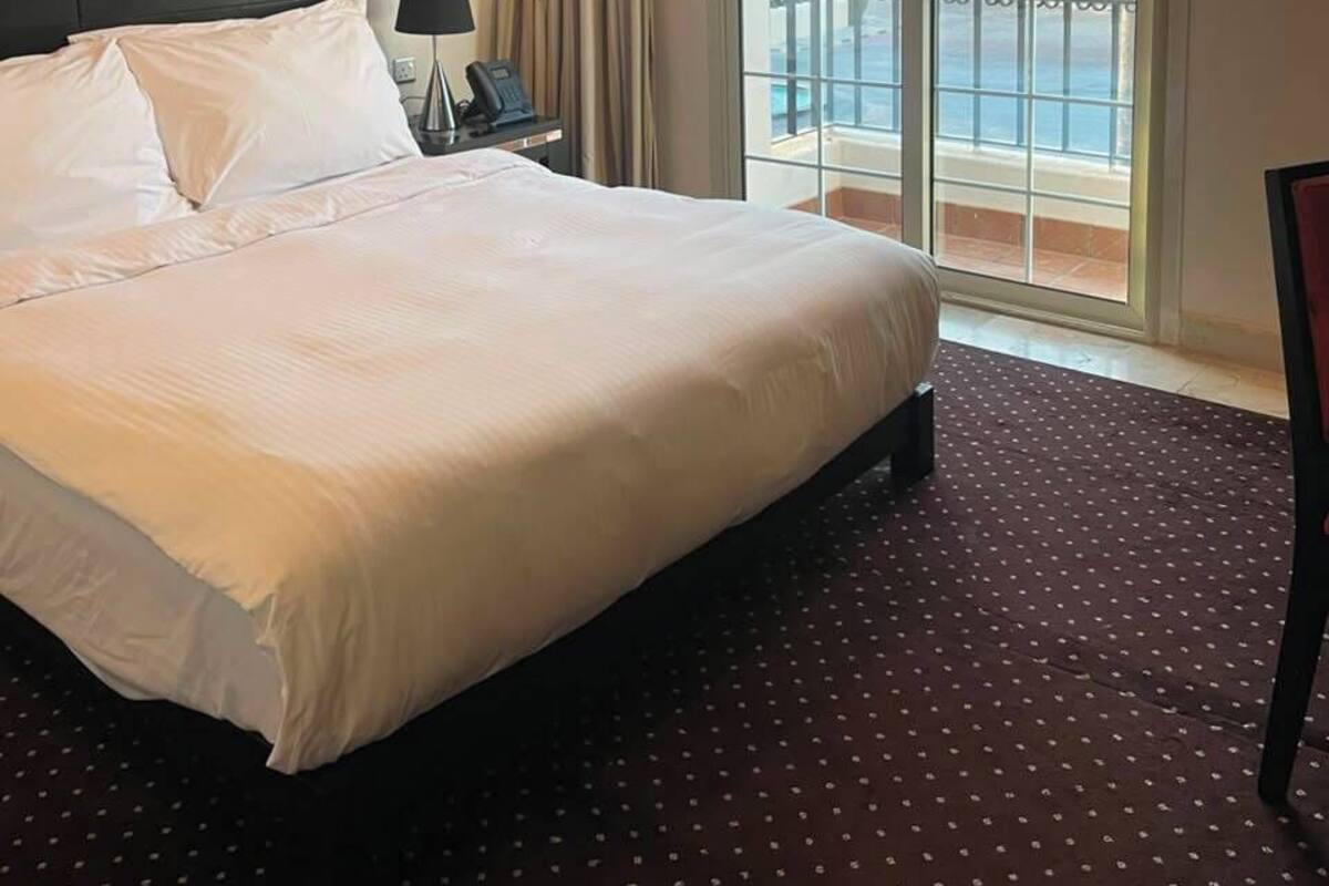 Ein Hotelzimmer mit einem großen Bett, einem Nachttisch, einer Lampe und Blick auf einen Pool und Palmen durch eine Glastür.