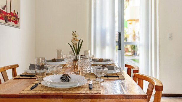 A wooden dining table set with glassware, plates, and cutlery, with a view of a garden through a glass door. - Mariscal Ideal Family Retreat, King Bed, Complete Linens, 220m from the Sea (Bombinhas)