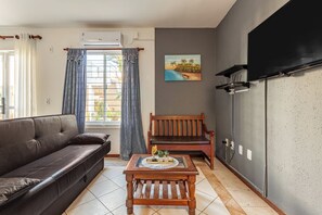 A living room with a sofa, a wooden coffee table, a bench, a painting, and a TV mounted on the wall. - Your home in Mariscal: Air conditioning and king-size bed 220m from the sea. (Bombinhas)