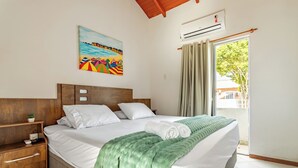 A neatly made bed with a green blanket, a wooden nightstand with a plant, a painting of a beach scene, and a window with curtains. - Mariscal Ideal Family Retreat, King Bed, Complete Linens, 220m from the Sea (Bombinhas)