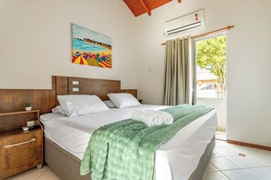 A neatly made bed with a green blanket, a wooden nightstand with a plant, a painting of a beach scene, and a window with curtains. - Your home in Mariscal: Air conditioning and king-size bed 220m from the sea. (Bombinhas)