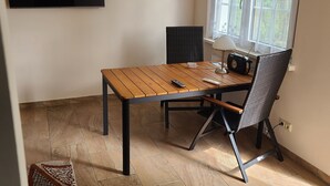 A room with a wooden table, a black chair, a television mounted on the wall, and a window with curtains. - Bungalow with lake view & fenced garden - quiet & private (Rüdersdorf bei Berlin)