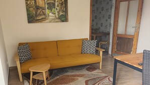 A living room with a sofa, a small table, a patterned rug, a painting on the wall, and a door with a patterned wallpaper. - Bungalow with lake view & fenced garden - quiet & private (Rüdersdorf bei Berlin)