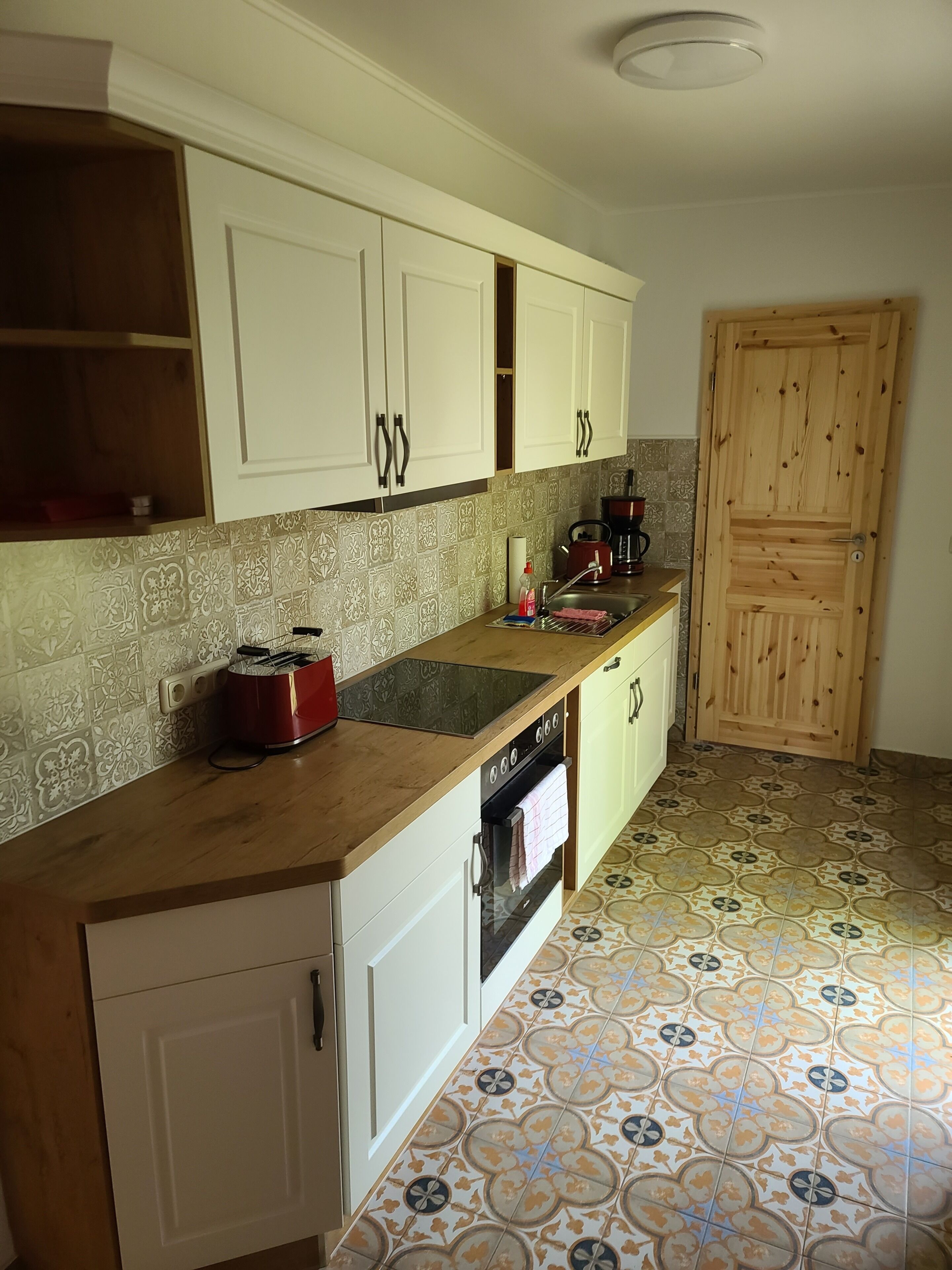 Une cuisine avec des meubles blancs, un plan de travail en bois, une bouilloire rouge, une machine à café et des carreaux de sol à motifs.
