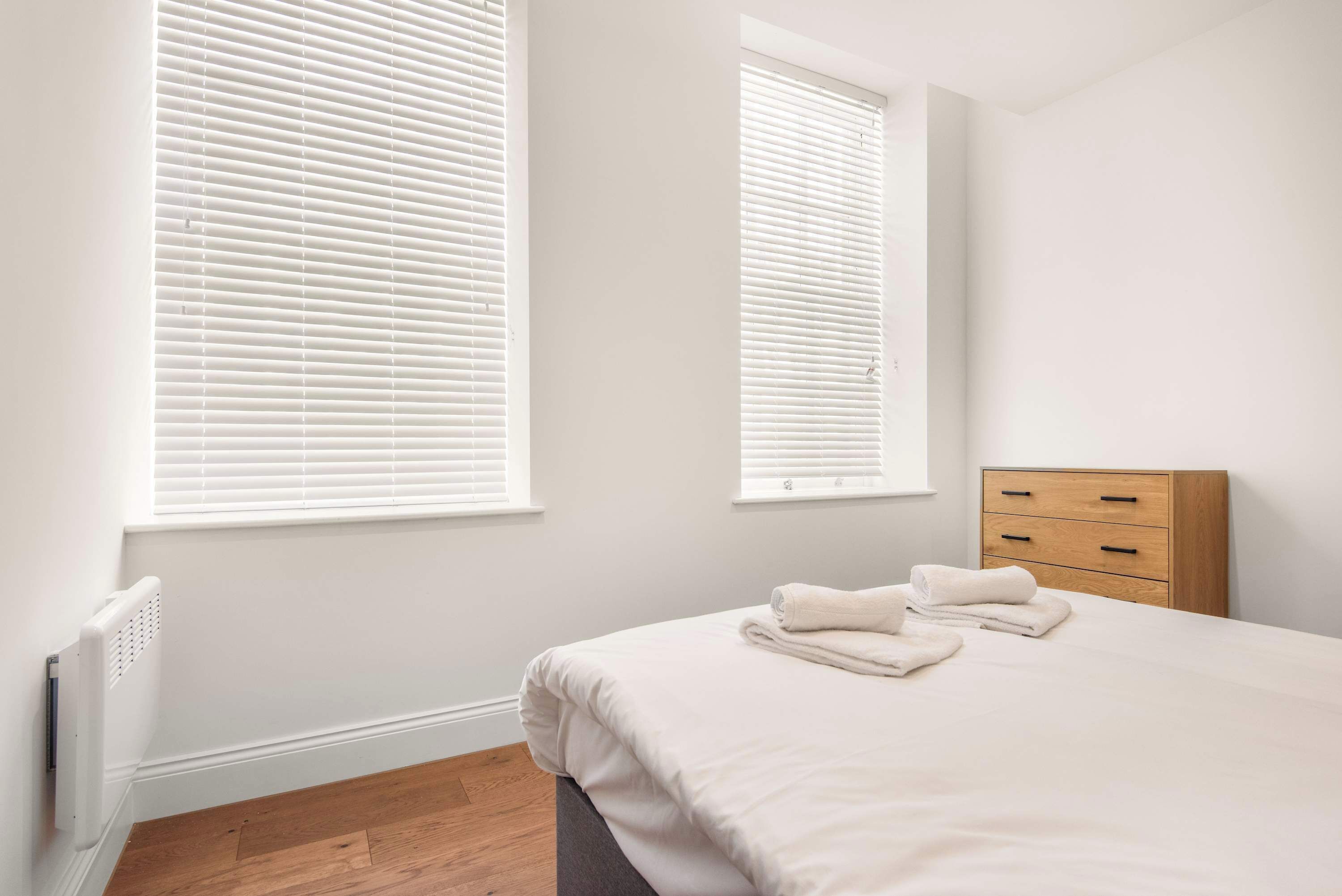 Un dormitorio con una cama, dos ventanas con persianas, un radiador y un aparador de madera.