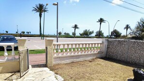 Une zone pavée, avec un mur en pierre, un portail en métal et une rue bordée de palmiers, où se trouve une voiture.