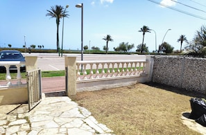 A paved area with a stone wall, a metal gate, and a street with palm trees and a car. - Fantástica casa a primera linea del mar en empuriabrava (Empuriabrava)