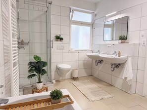 Un baño moderno con cabina de ducha de vidrio, azulejos blancos, un espejo grande, dos lavamanos y una planta en maceta.