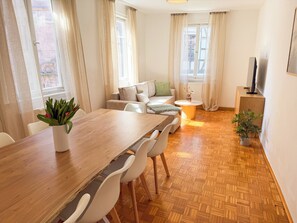 A modern living room with a wooden dining table, white chairs, a sofa, and a TV on a wooden cabinet. - 140 sqm apartment for 6–10 people, large living/dining area & garage (Gernsbach)