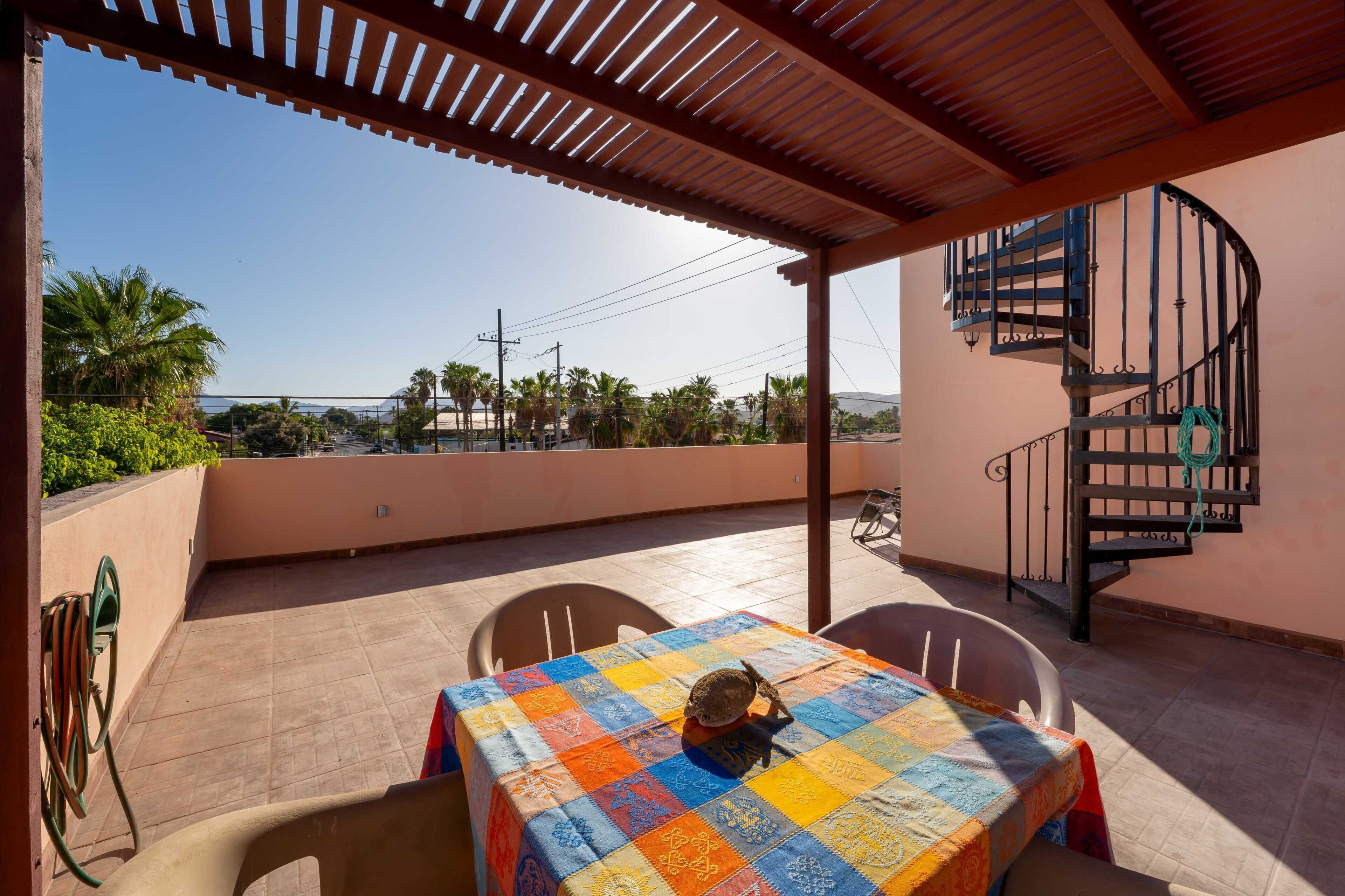 A covered outdoor patio with a table and chairs, a spiral staircase, and a view of palm trees and distant mountains.