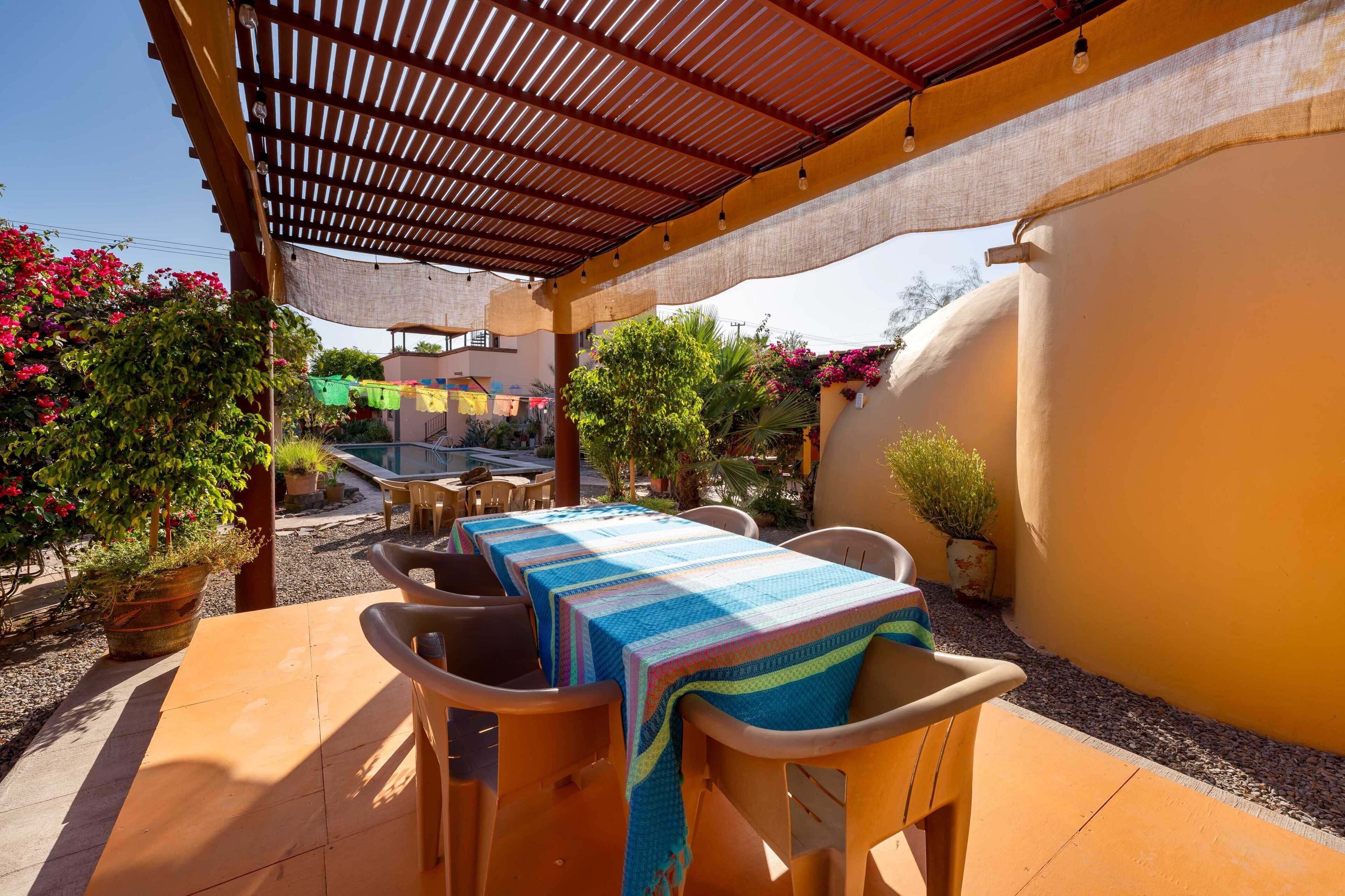 Un patio al aire libre con mesa y sillas, cubierto por una pérgola, rodeado de plantas en macetas y una piscina.