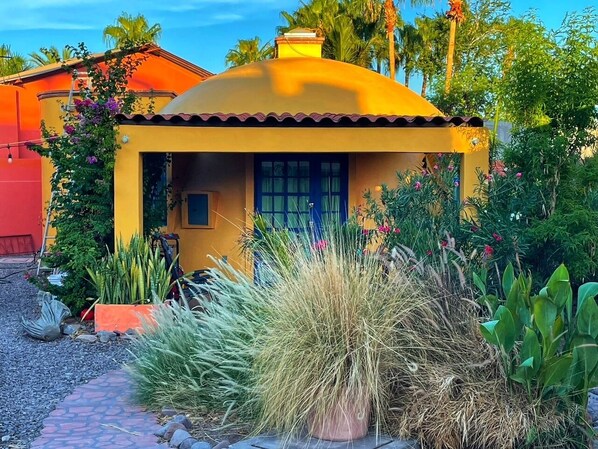 Una casa amarilla con techo rojo, rodeada de follaje y flores.