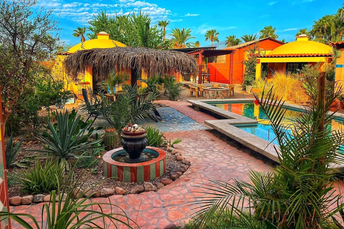 Un área de piscina con una estructura de techo de paja, una zona de comedor y una fuente de agua.