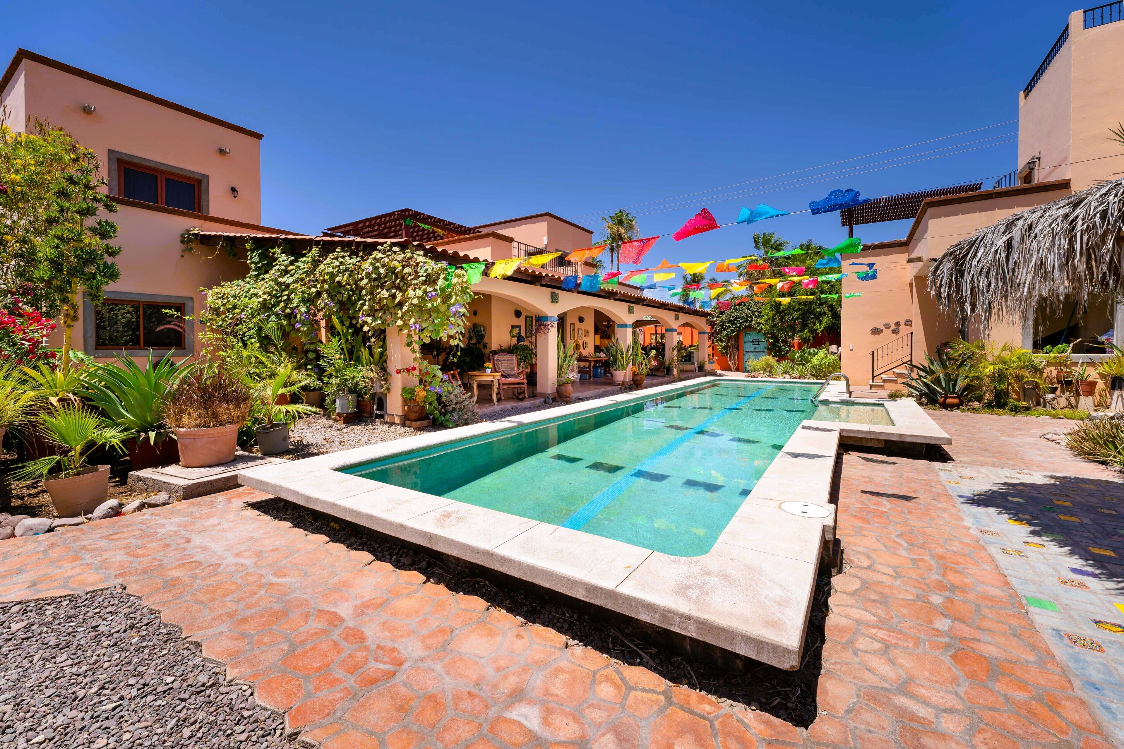 Una piscina con un patio de piedra, rodeada de plantas en macetas y un edificio con balcón.