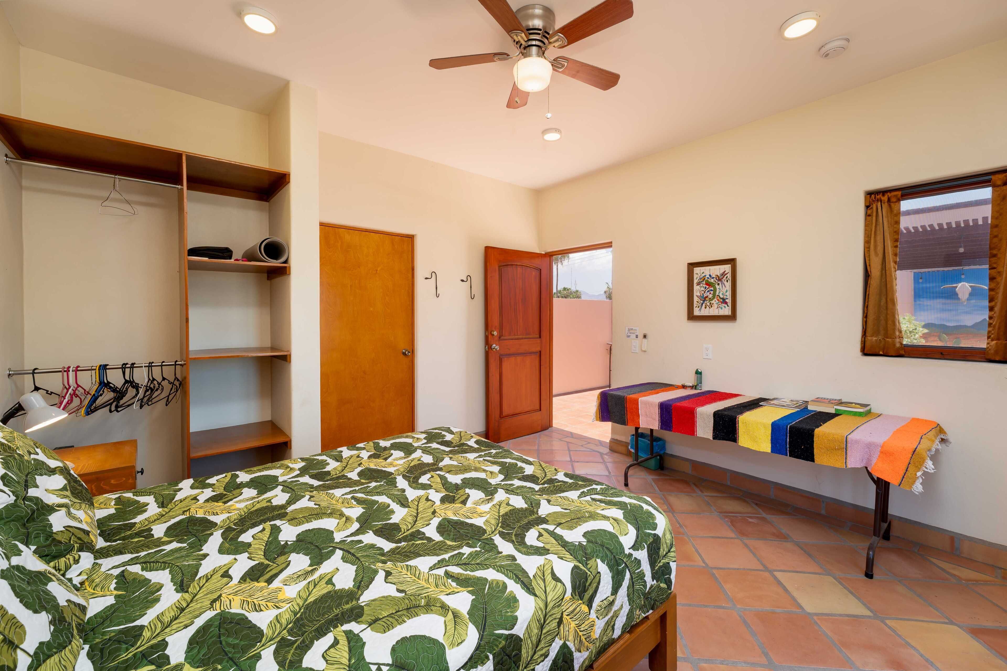 A bedroom with a bed, a wardrobe, a ceiling fan, a window with curtains, and a table with a colorful tablecloth.