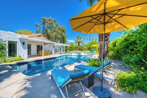Une piscine avec des chaises longues et un parasol, entourée de verdure et d’une maison.