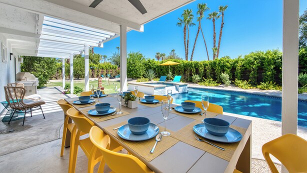 An outdoor dining area with a pool view, yellow chairs, and a ceiling fan. - Sun and Games -  - The Ultimate Central Palm Springs Escape! (Palm Springs)