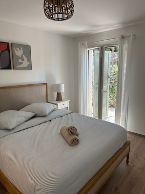 A bedroom with a bed, a nightstand, a lamp, a wall art piece, and a view through a window with shutters. - Villa Rosa By Avecoeur (Sainte-Maxime)