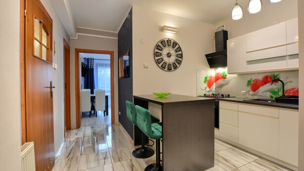 A modern kitchen with a central island, a wall clock, and a dining area with a chair. - Art Apartament Krynica Zdrój Centrum (Krynica-Zdrój)