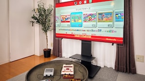Ein Nintendo Family Computer mit Bildschirm für die Spielauswahl, ein kleiner Tisch mit Retro-Spielkonsolen und eine Topfpflanze in einem Raum mit schlichter Wand und Vorhängen.