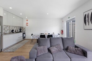Un soggiorno moderno con un divano grigio, un'area pranzo con tovaglia bianca e una cucina con mobili bianchi.