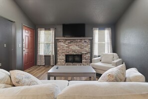 A living room with a brick fireplace, a flat-screen TV mounted above it, and a sofa with patterned cushions. - The Red Door on 66th - 3BR/2BA Duplex (Tulsa)