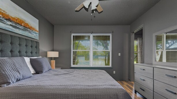 A bedroom with a large bed, a ceiling fan, a window with blinds, a dresser, and a painting on the wall.