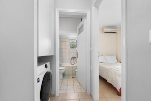 A narrow hallway leading to a bathroom with a toilet, shower, and a bed with a white bedspread. - Abatis CityHome (Lefkada)