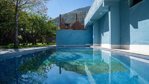 Piscina cristalina com borda azulejada, rodeada por um edifício de paredes azuis e uma montanha ao fundo.