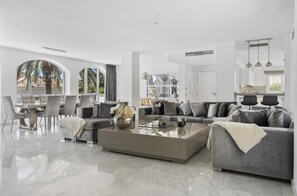 A spacious living area with a large coffee table, grey sofas, and a dining area with chairs. - Villa Elviria (Marbella)