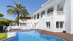 A pool with a wooden deck, a white building with arched windows, and a palm tree. - Villa Elviria (Marbella)