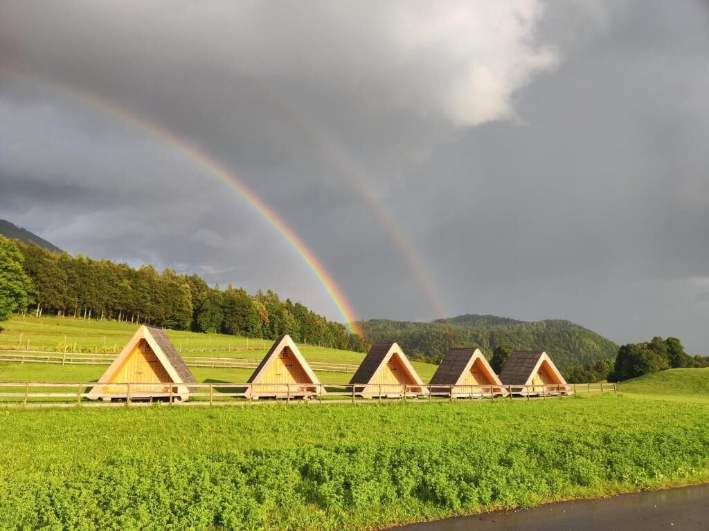 A row of wooden A-frame cabins in a green field with a rainbow in the sky.