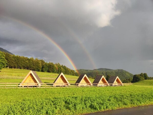 A row of wooden A-frame cabins in a green field with a rainbow in the sky.