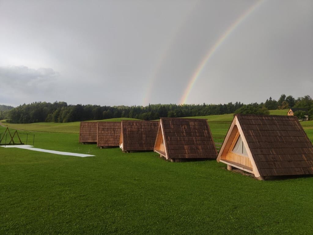 Una hilera de estructuras con techo de madera en un campo cubierto de hierba, con un arcoíris al fondo.