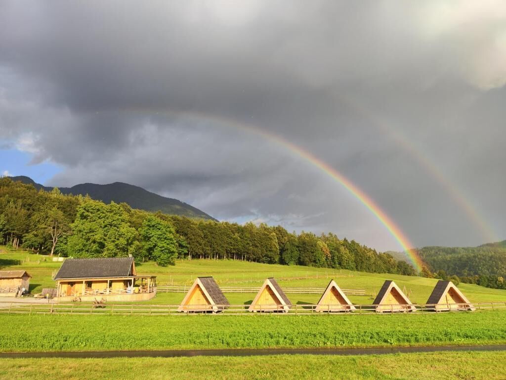 Filetta di cabine con tetto a punta in un prato erboso, con montagne sullo sfondo e un arcobaleno.
