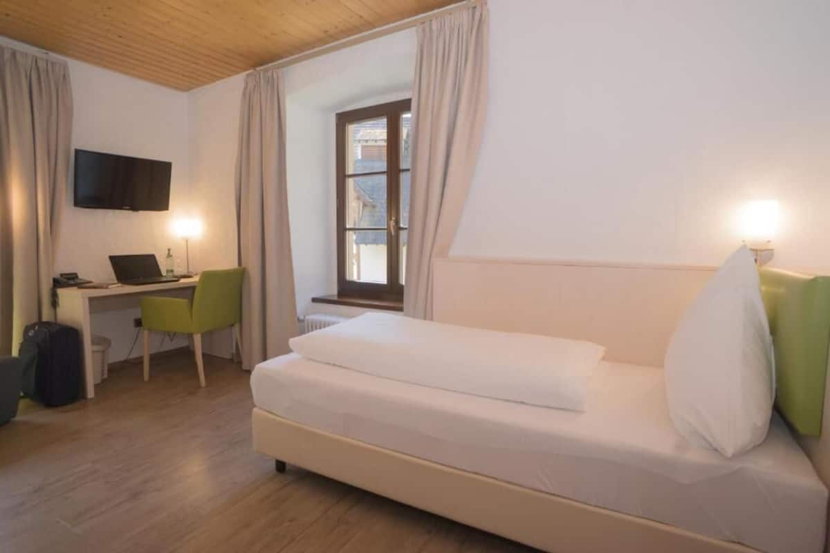 A hotel room with a bed, a desk with a computer, a chair, a TV mounted on the wall, and a window with curtains.