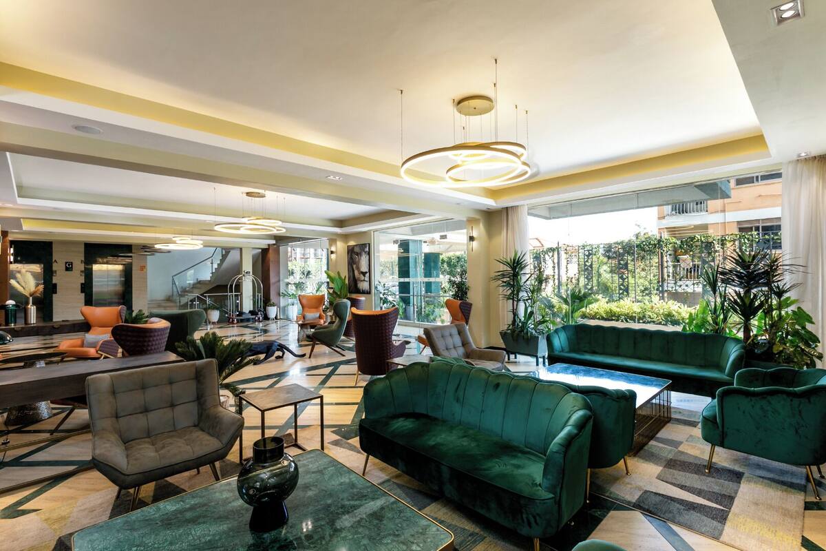 Eine großzügige Lobby mit einem grünen Sofa, Sesseln und einem Couchtisch. Ücht Topfpflanzen und ein großes Fenster bieten Ausblick ins Freie.