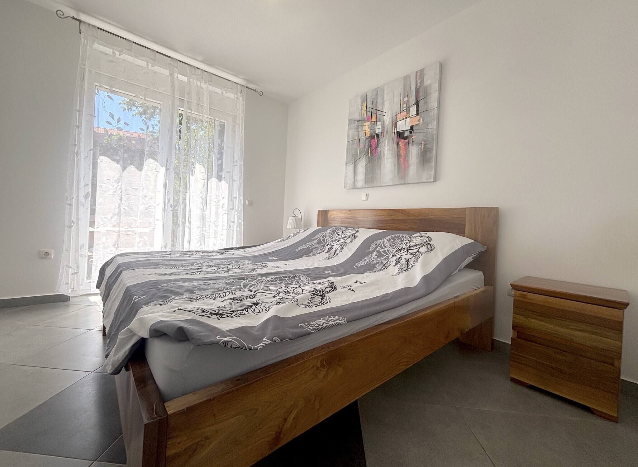 A bedroom with a wooden bed, a nightstand, a window with sheer curtains, and a wall-mounted abstract painting.