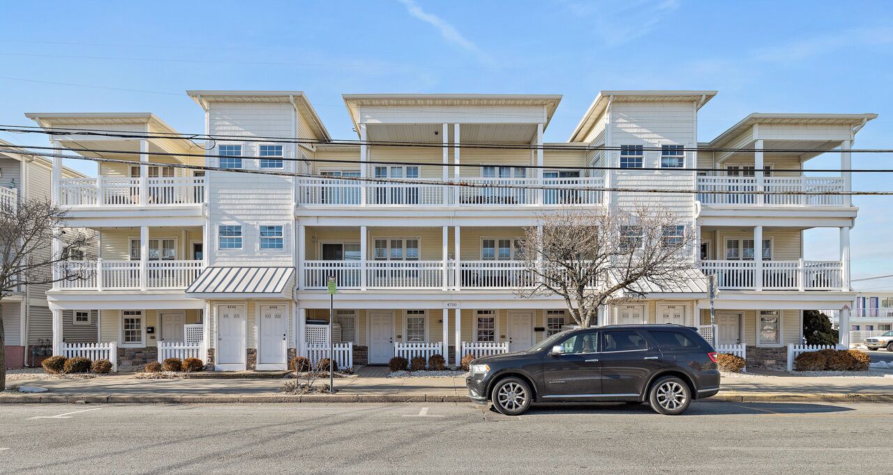 A multi-story residential building with balconies, a car parked in front, and a clear sky.