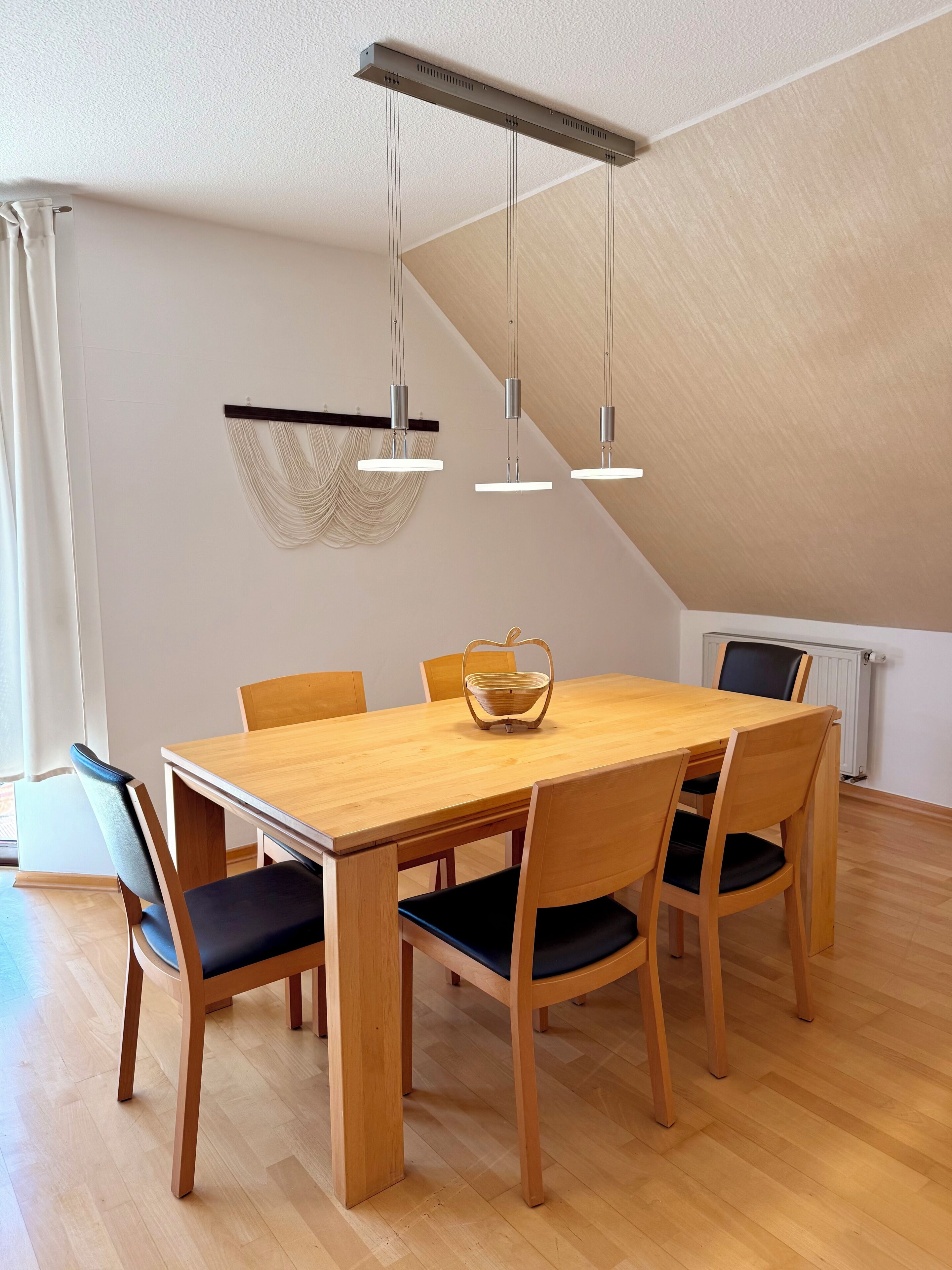 A dining area with a wooden table, six chairs, a hanging light fixture, and a woven wall decoration.