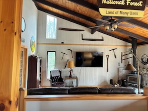 A rustic cabin interior with a wooden ceiling, a television, a fireplace, and various tools on the walls.