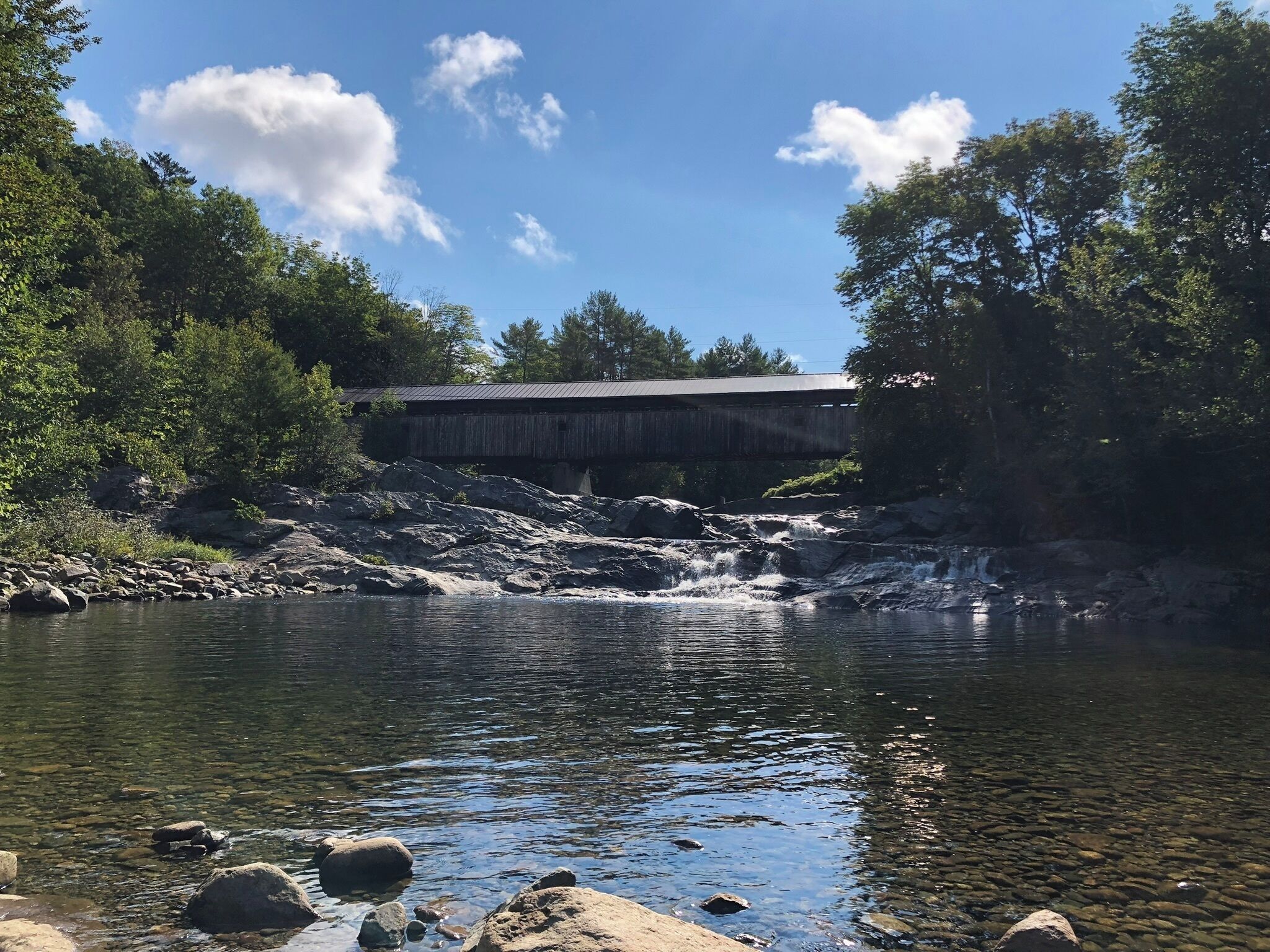 A covered bridge over a river with clear water, surrounded by trees and rocks.
