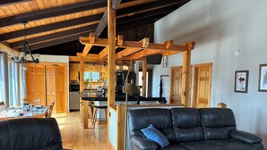 Un'ampia zona giorno con un divano in pelle nera, travi in legno a soffitto, un tavolo da pranzo e una cucina sullo sfondo.