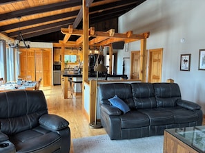 A spacious living area with a black leather sofa, wooden ceiling beams, a dining table, and a kitchen in the background.