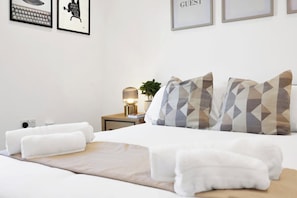 Cama bem arrumada com lençóis brancos, luminária de cabeceira e almofadas com padrão geométrico.