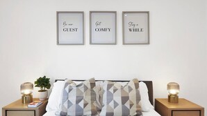 Cama bem arrumada com almofadas estampadas, citações motivacionais emolduradas na parede e luminárias de cabeceira sobre mesas de cabeceira de madeira.