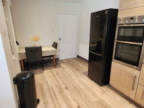 A compact kitchen with a refrigerator, oven, and a dining table with chairs. - 8 Arvonia Terrace (Criccieth)